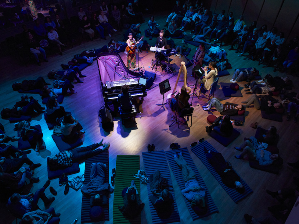 Musicians perform onstage under colorful lighting as audience members recline on mats during a live concert.