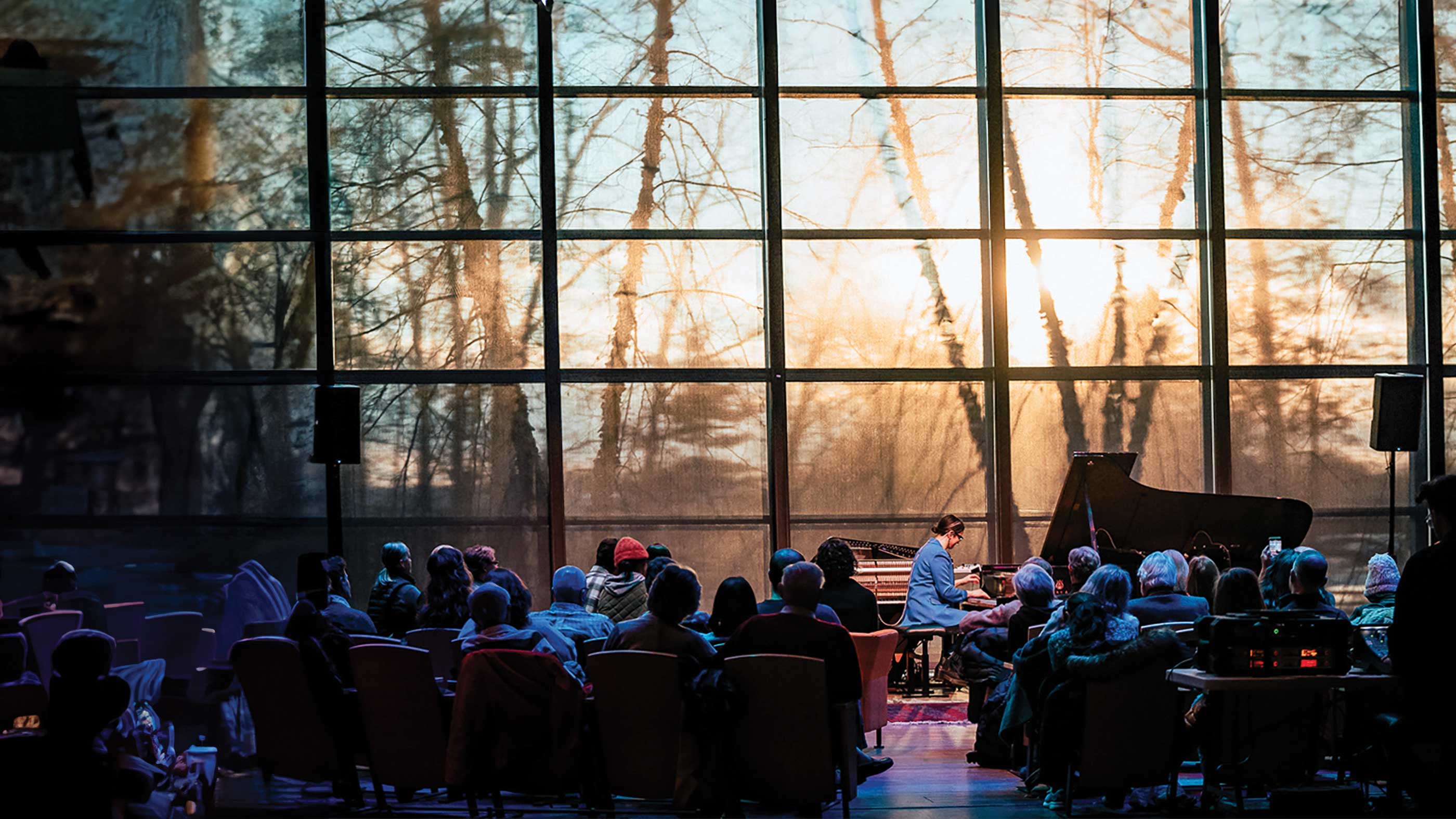 A pianist performs for an audience inside Zankel Music Center, with sunlight and tree silhouettes visible through the tall glass wall behind the stage.