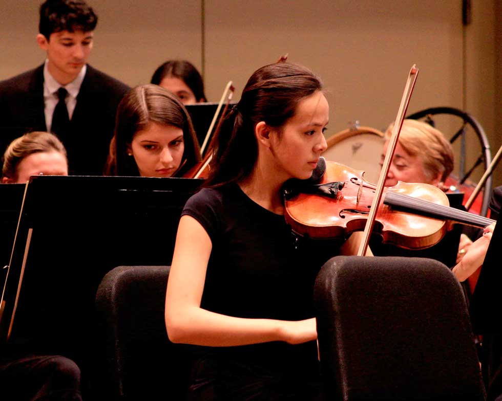 A student violinist performs with an orchestra, with other musicians seated behind her.