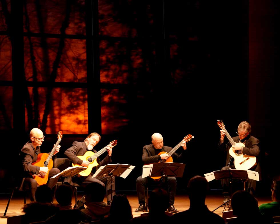 Four guitarists perform onstage at Zankel Music Center under warm concert lighting.