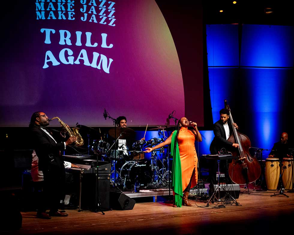 A jazz ensemble performs “Trill Again” onstage with a vocalist, saxophonist, drummer, bassist, and percussionist.