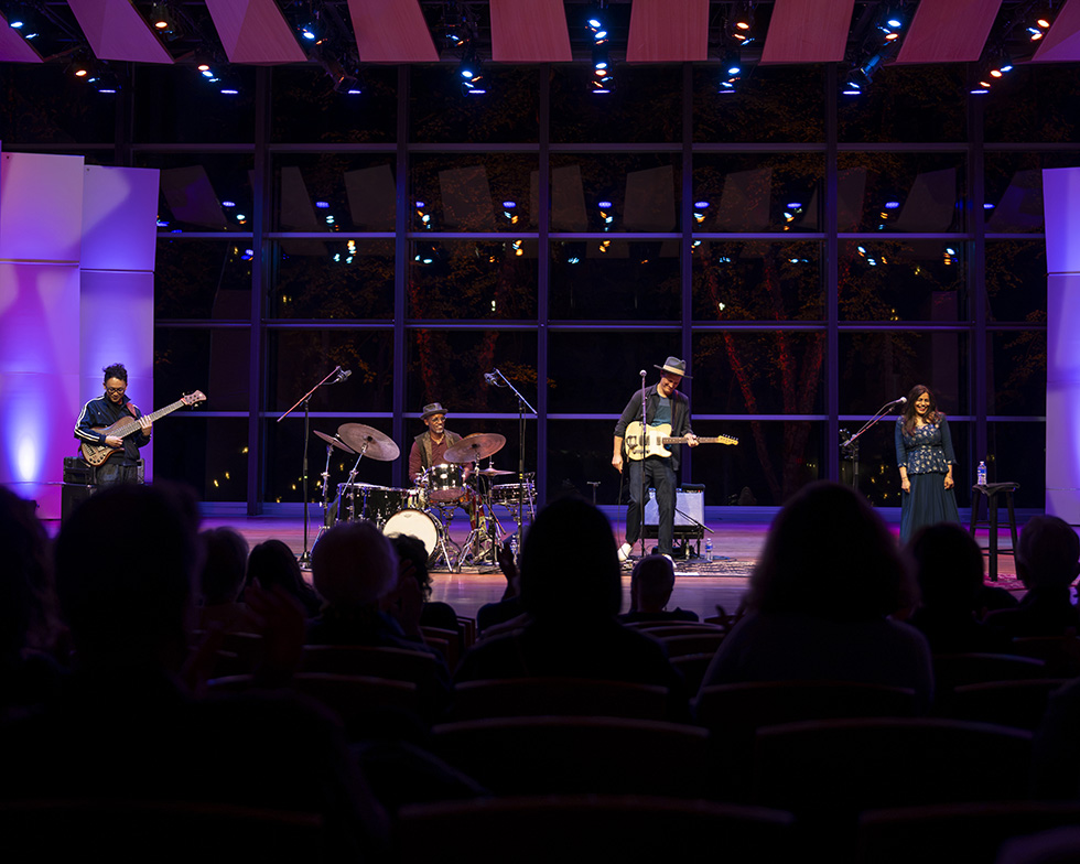 Four musicians perform onstage with guitars, drums, and vocals under colorful lighting as an audience watches.